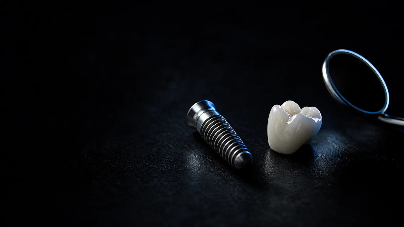 A titanium implant, porcelain crown and dental mirror rest under cool light on a dark clinical surface.